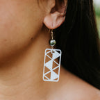 A close-up of a person's ear wearing a white geometric cutout rectangular dangle earring with a small dark bead and gold hook, next to a yellow flower in her hair.