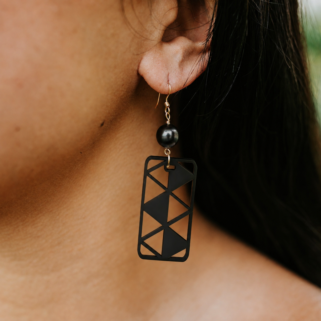 A close-up of a person's ear wearing a black geometric rectangular drop earring with a small black bead and gold-tone hook.