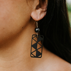A close-up of a person's ear wearing a black geometric rectangular drop earring with a small black bead and gold-tone hook.