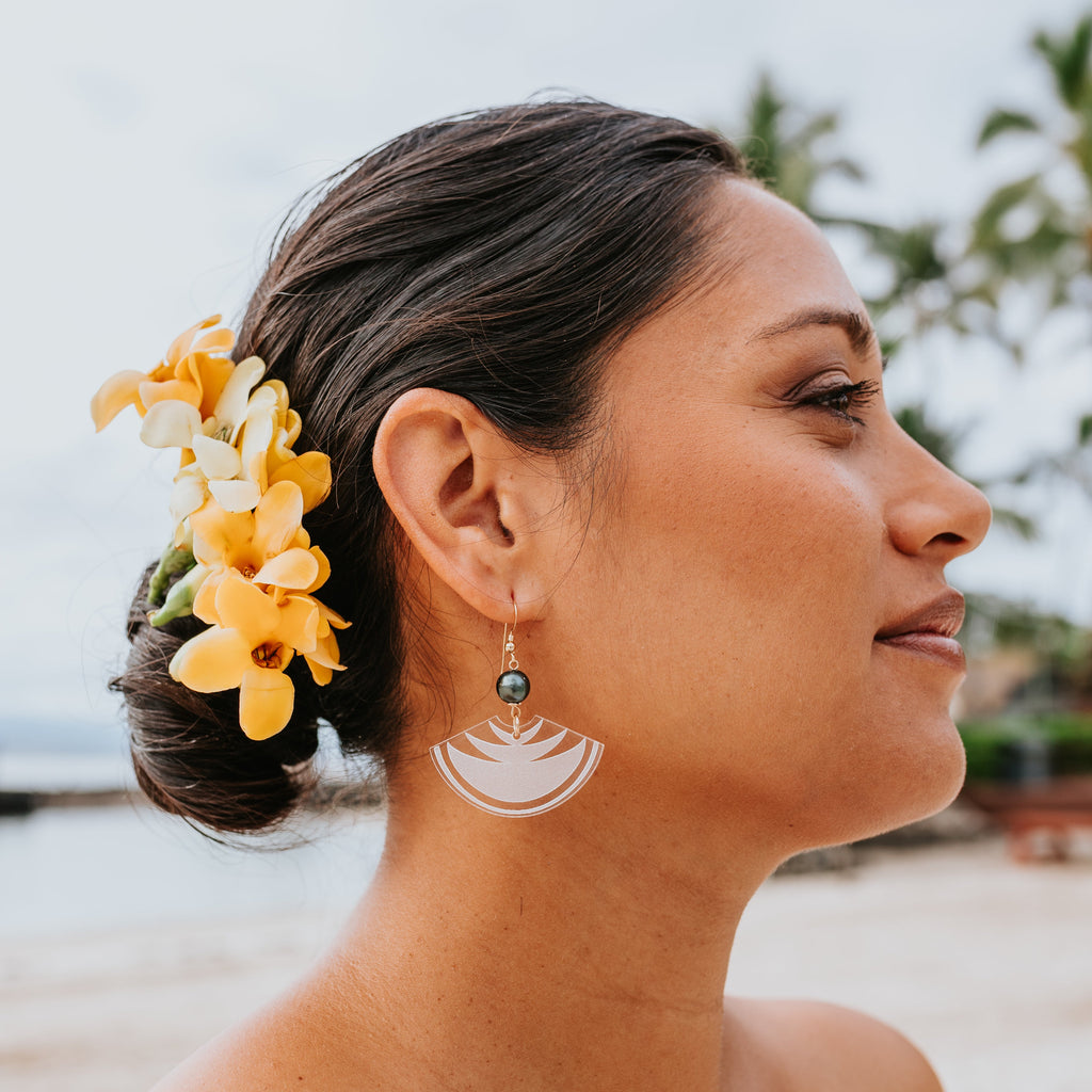 A woman in profile at a beach wearing a white semicircular acrylic dangle earring with a small dark green bead, with yellow and white plumeria flowers in her hair.