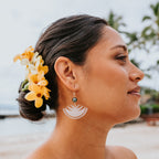 A woman in profile at a beach wearing a white semicircular acrylic dangle earring with a small dark green bead, with yellow and white plumeria flowers in her hair.