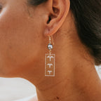 A close-up side profile of a woman wearing a delicate silver dangle earring. The earring features a small round silver bead and a rectangular silver frame with three cutout birds flying upwards.