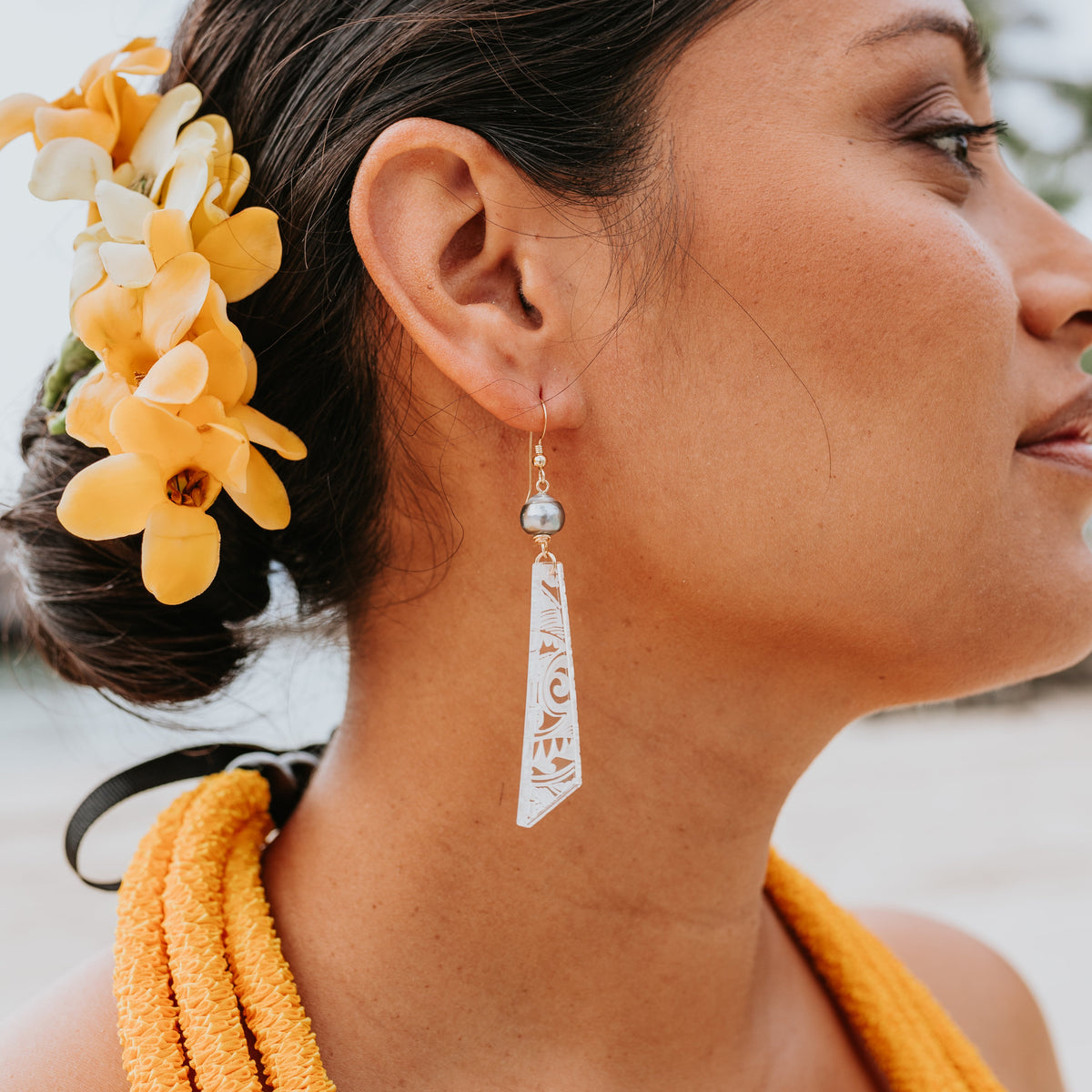 A close-up side profile of a woman wearing a long, silver-toned drop earring with engraved text, styled with yellow and white flowers in her hair and a yellow necklace.